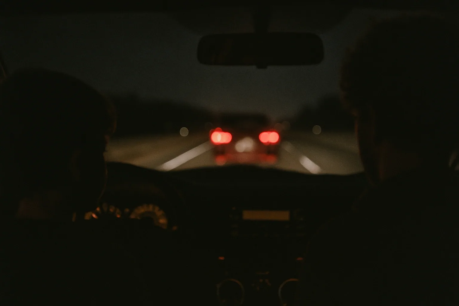 A dark highway stretches ahead through a windshield at night, taillights visible and dashboard glow low.